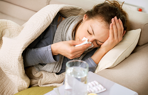 Woman laying on a couch in a blanket and blowing her nose