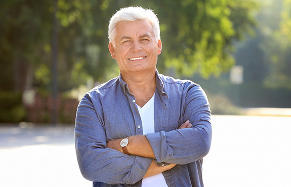Older man in a gray button down shirt smiling with his arms crossed